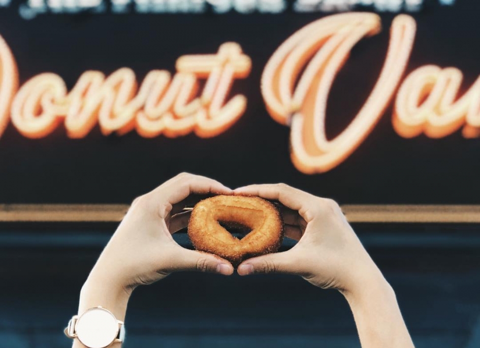 Two hands framing a fresh cinnamon donut in front of the van's orange script sign