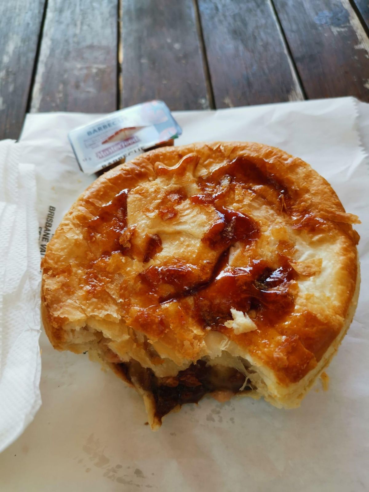 A golden Fernvale pie on its paper bag, with a barbecue sauce tub, on a timber picnic table