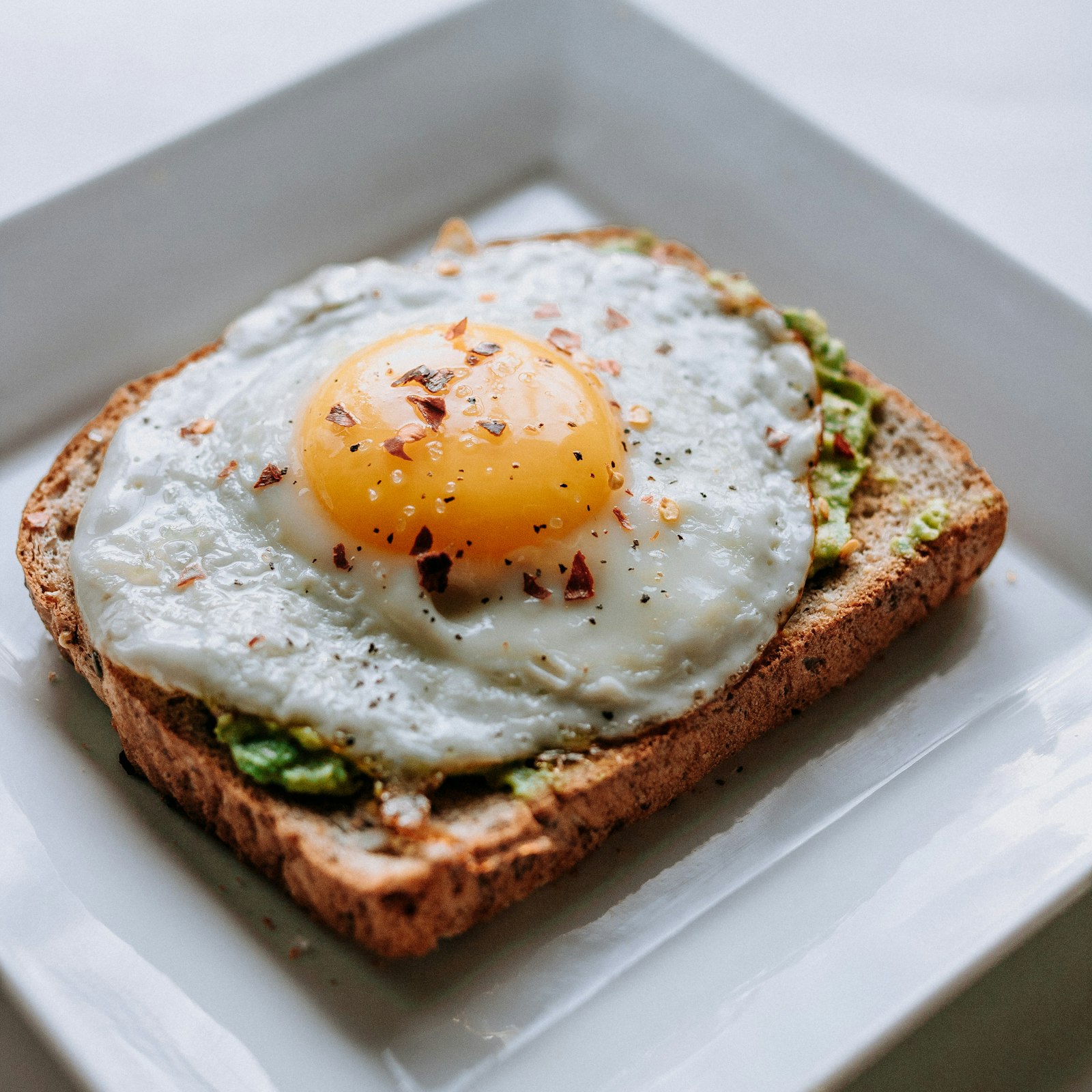 A fried egg on avocado toast, served on a square white plate